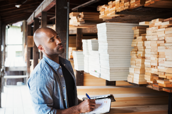 Man standing in a lumber yard, holding a folder, checking wood, learning what is MOQ, writing notes.